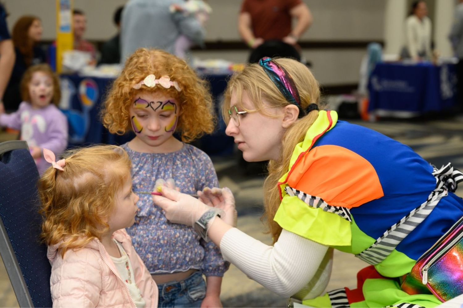 A girl has her face painted by a clown while her sister watches
